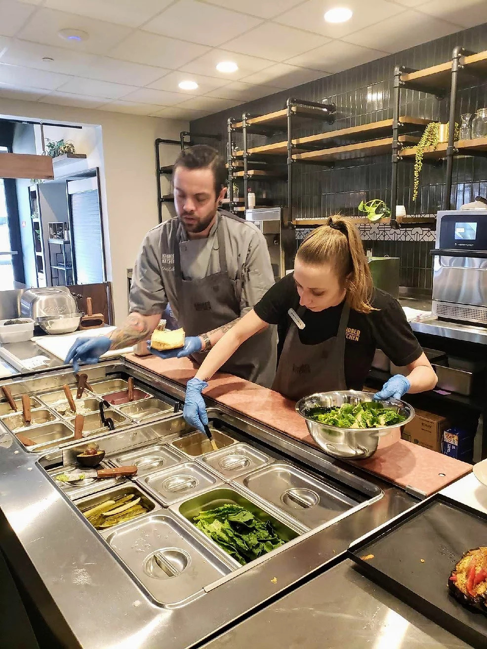Photo of a cafe employee and trainee preparing a bowl of salad ingredients.