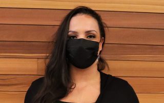 Photo of a woman in front of a wood wall, wearing a black face mask.