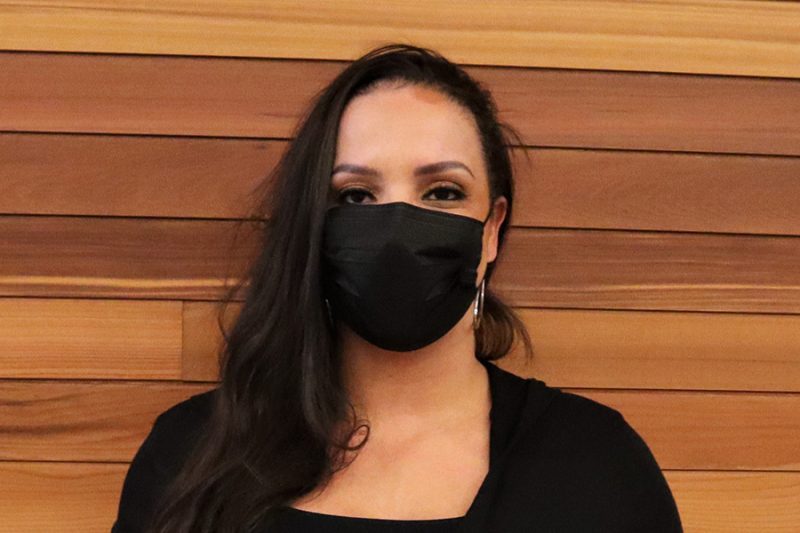 Photo of a woman in front of a wood wall, wearing a black face mask.
