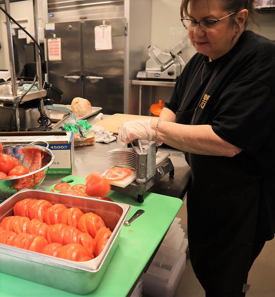A woman slicing tomatoes with a vegetable slicer in a commercial kitchen.