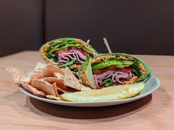 Photo of a bisected club wrap in a spinach tortilla, served with tortilla chips and a pickle on a plate.